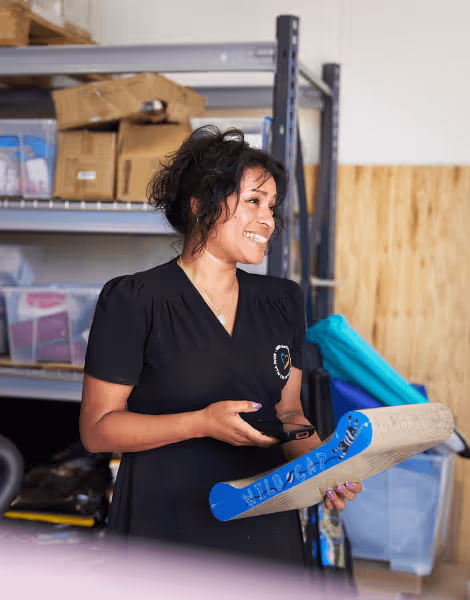 Bilden visar en leende kvinna med lockigt hår som håller en mobiltelefon och ett blått rör märkt med "Wildcat". Hon står i ett lager eller arbetsrum, omgiven av hyllor fulla med kartonger och diverse föremål.