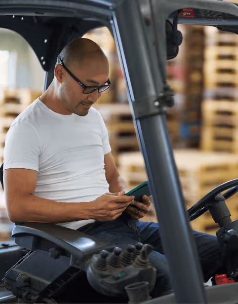 Bilden visar en man med glasögon som sitter i en gaffeltruck och använder en mobiltelefon. Han är klädd i en vit t-shirt och arbetar i en lageromgivning med pallar staplade i bakgrunden.