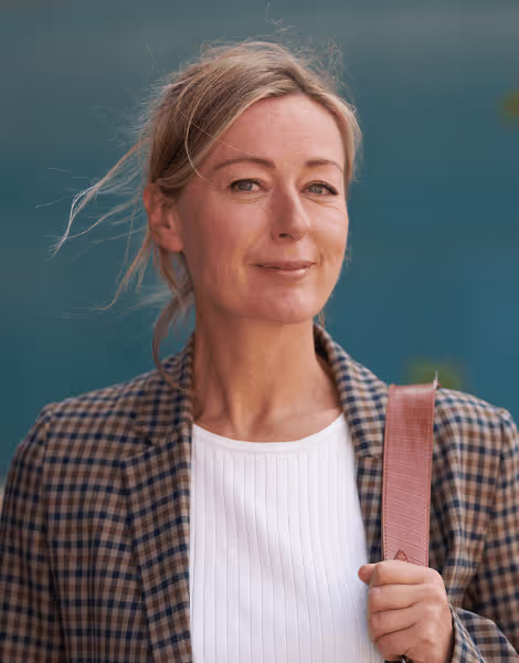 Bilden visar en kvinna med blont hår uppsatt i en lös knut, klädd i en rutig kavaj och en vit tröja. Hon ler mjukt mot kameran och har en väska över axeln i en utomhusmiljö med en suddig, blå bakgrund.