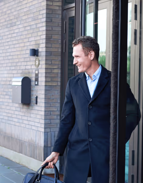 Bilden visar en leende man som kliver ut genom en dörr, klädd i en mörk kappa och blå skjorta. Han håller en läderväska i handen och befinner sig framför en tegelbyggnad.