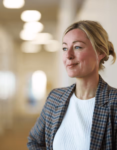 Bilden visar en kvinna med blont hår uppsatt, klädd i en rutig kavaj och en vit tröja, som står med ett mjukt leende och ser åt sidan. Hon befinner sig inomhus, i en upplyst korridor med suddiga lampor i bakgrunden.