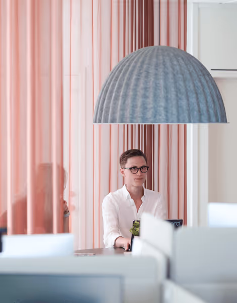 Bilden visar en man med glasögon som sitter vid ett skrivbord i ett modernt kontor, klädd i en ljus skjorta. Rummet har rosa draperier och en stor, rund lampa som hänger ovanför honom, med suddiga konturer av en annan person i bakgrunden.