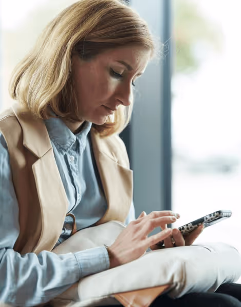 Bilden visar en kvinna med blont hår som sitter och använder sin smartphone. Hon är klädd i en ljusblå skjorta och en beige väst, och verkar vara koncentrerad på skärmen framför henne.