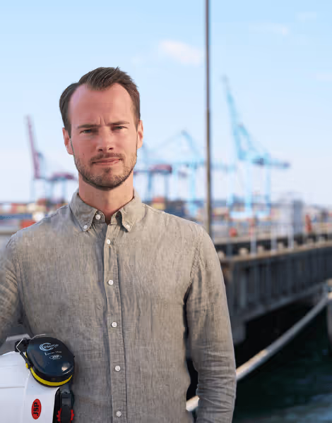 Bilden visar en man i ljus skjorta som håller en vit skyddshjälm med hörselskydd vid kajen i en hamn. Bakom honom syns flera kranar och containrar, vilket skapar en industriell bakgrund mot en blå himmel.