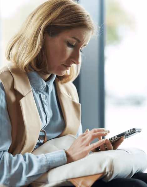 Bilden visar en kvinna med blont hår som sitter och använder sin smartphone. Hon är klädd i en ljusblå skjorta och en beige väst, och verkar vara koncentrerad på skärmen framför henne.