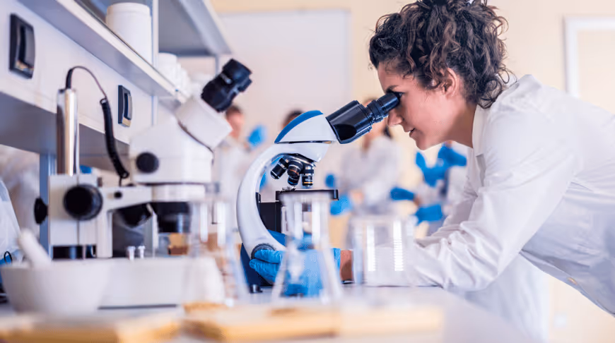 Forskare i vit labbrock tittar i ett mikroskop i ett laboratorium med kollegor i blå handskar i bakgrunden.