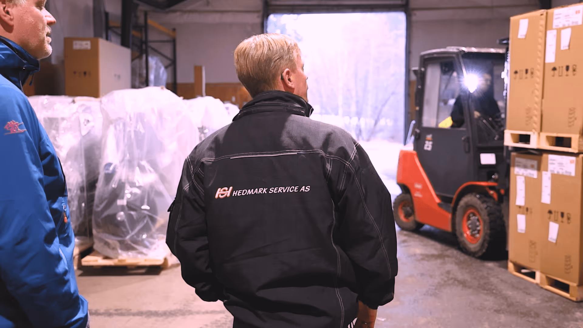Två män i arbetskläder i ett lagerutrymme där en gaffeltruck flyttar pallar med inplastat gods och kartonger.