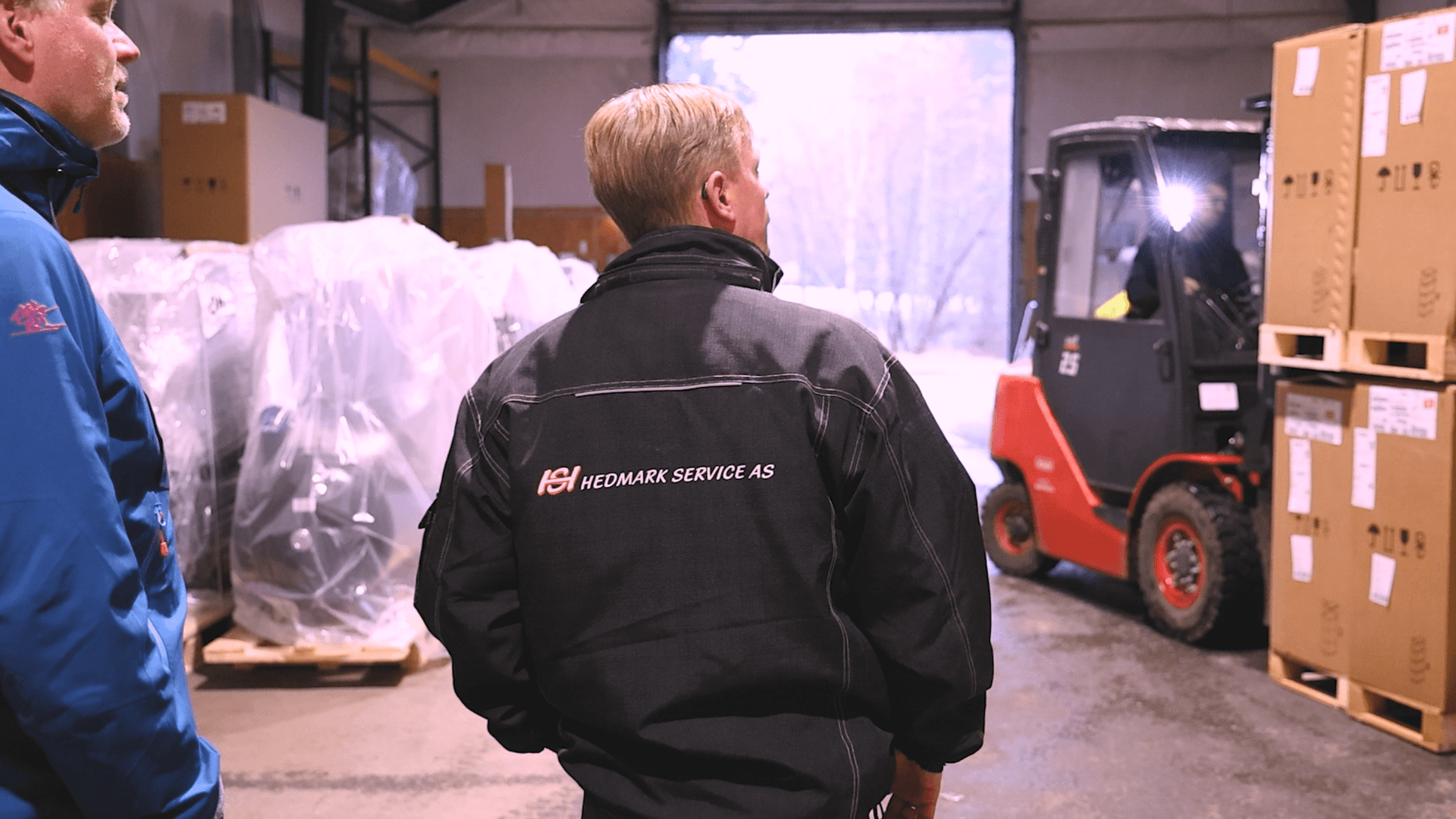 Två män i arbetskläder i ett lagerutrymme där en gaffeltruck flyttar pallar med inplastat gods och kartonger.