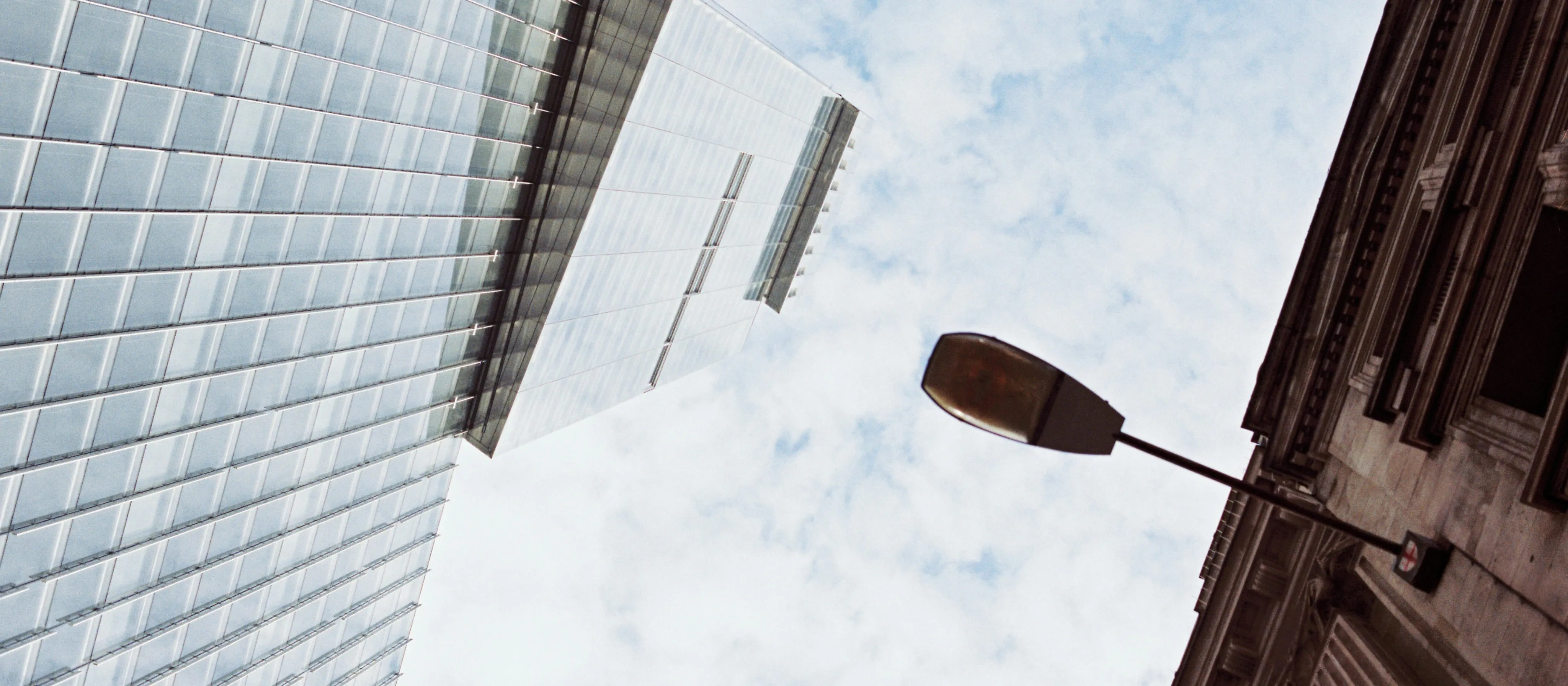 An undershot image of a modern glass building next to an older decorative building and streetlight.