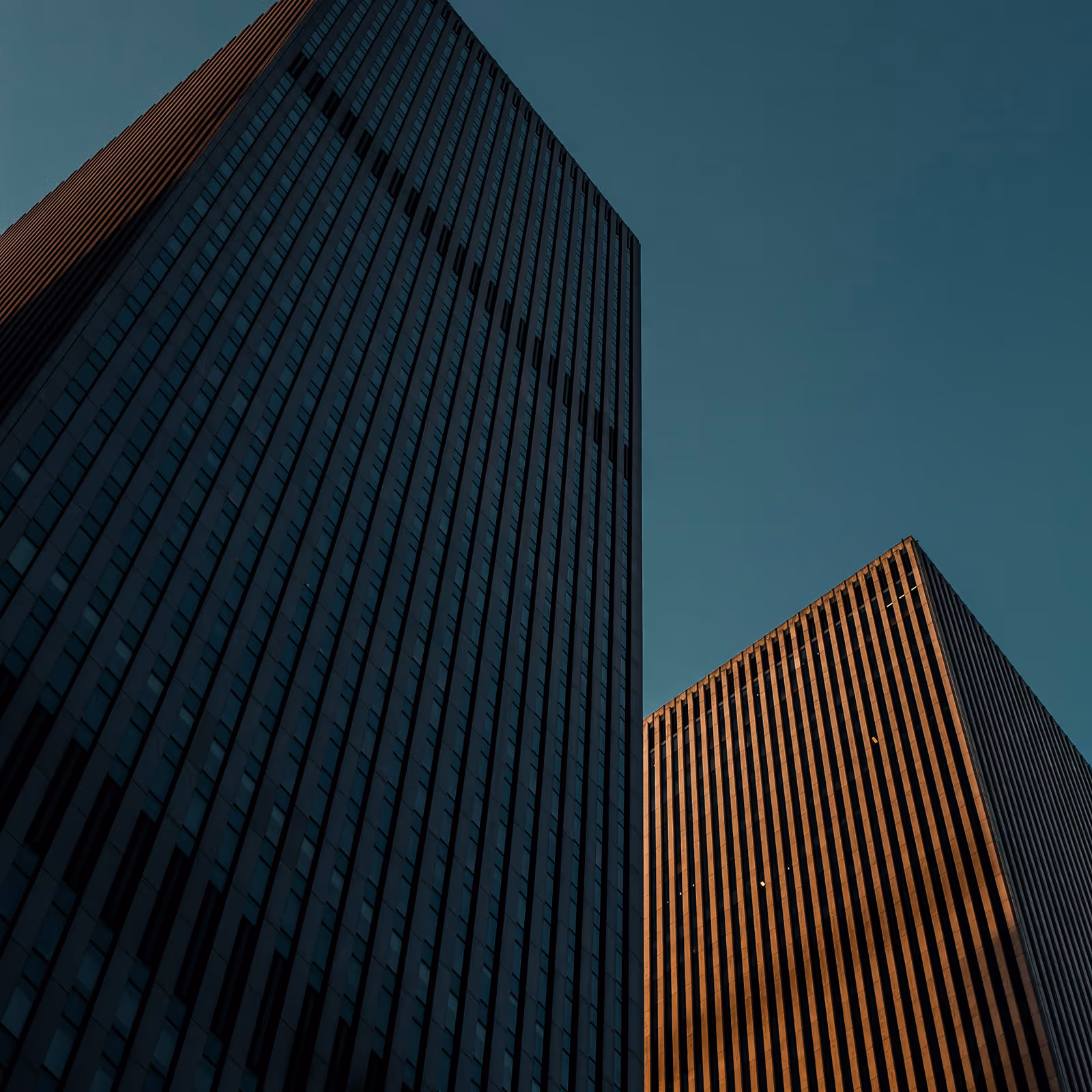 Two tall modern skyscrapers with vertical lines against a clear blue sky during sunset.