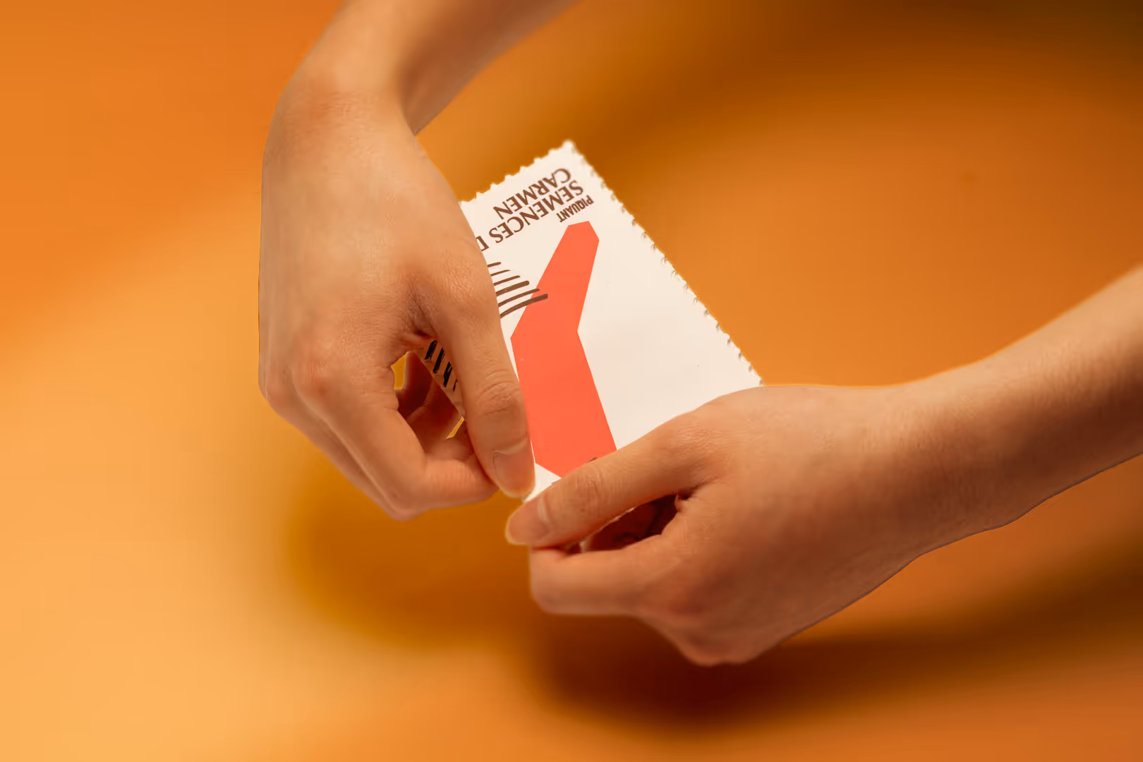 Hands carefully holding a red seed packet with perforated stamp edges on an orange background.