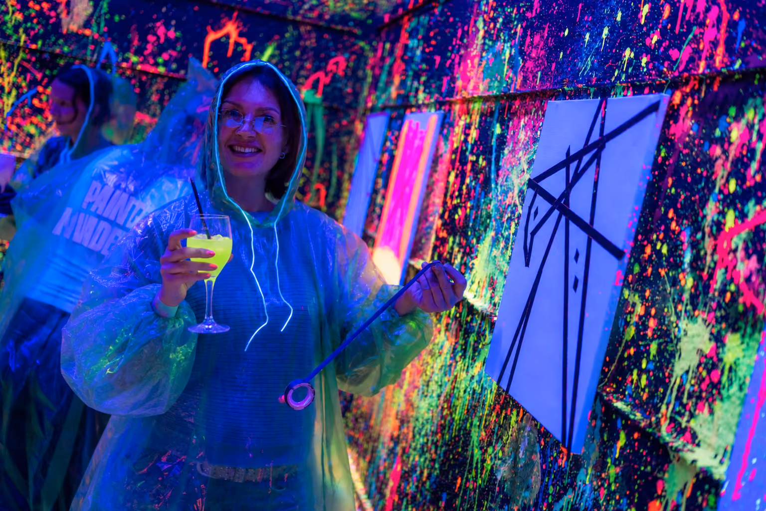 Une femme souriante portant un poncho transparent est en train de peindre une toile blanche avec des bandes noires abstraites dans une pièce décorée de peinture fluorescente colorée sous lumière noire, elle tient un verre avec un liquide jaune vif.