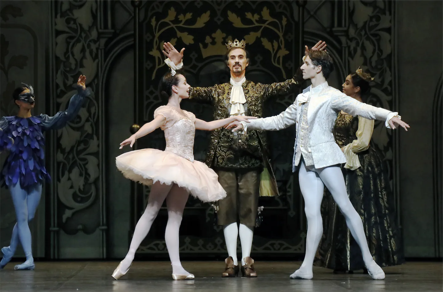 Quatre danseurs de ballet en costume d'époque sur scène, avec deux danseurs classiques au centre tenant les mains, un homme couronné derrière eux et d'autres personnages en arrière-plan.