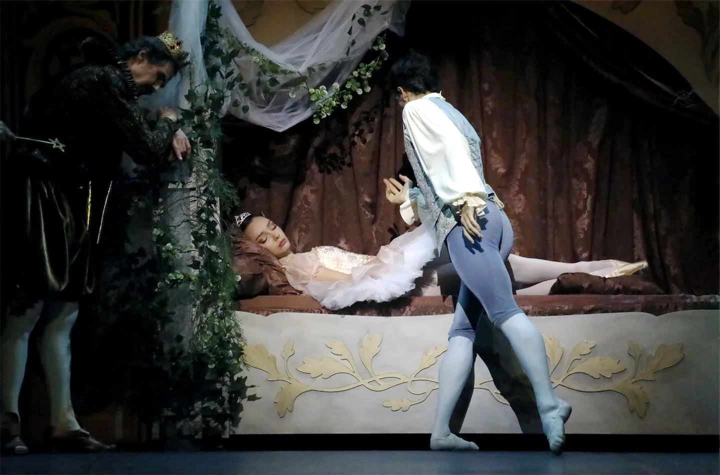 Une danseuse en tutu blanc et diadème est endormie sur un lit orné, un danseur en costume classique s'approche, et un homme vêtu de noir avec une couronne observe à côté.