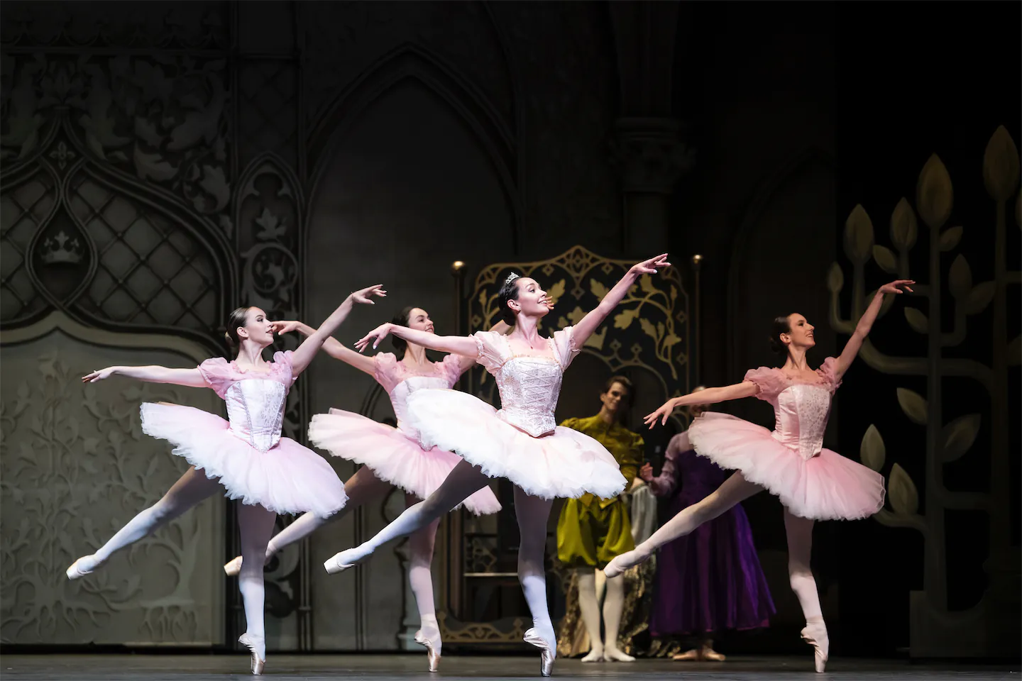 Quatre danseuses en tutu rose exécutant une pose de ballet classique sur scène avec un décor gothique en arrière-plan.