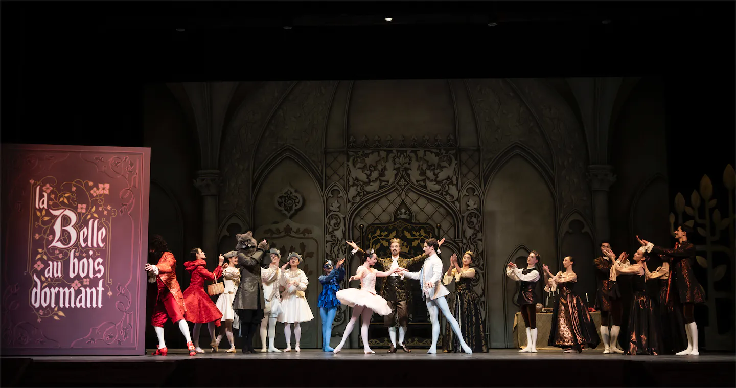 Scène de ballet avec des danseurs en costumes d'époque sur scène devant un décor gothique, accompagnés d'un panneau indiquant ‘la Belle au bois dormant’.