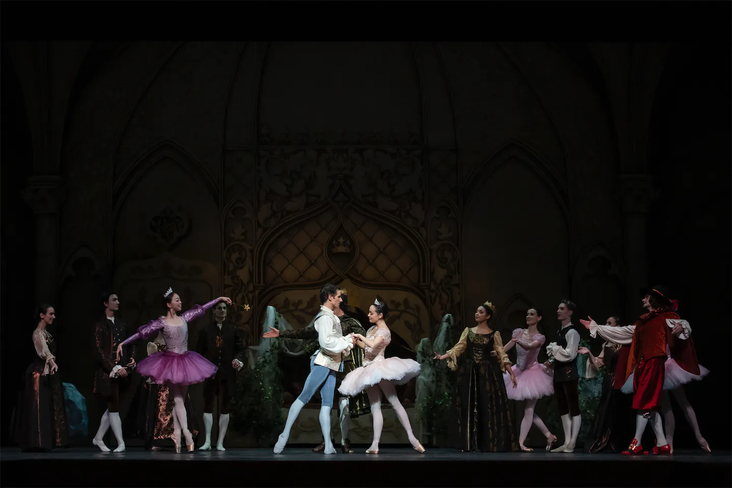 Danseurs de ballet en costume sur scène avec un décor de château médiéval, la danseuse centrale en tutu rose pâle et le danseur en collants bleus se tiennent la main.
