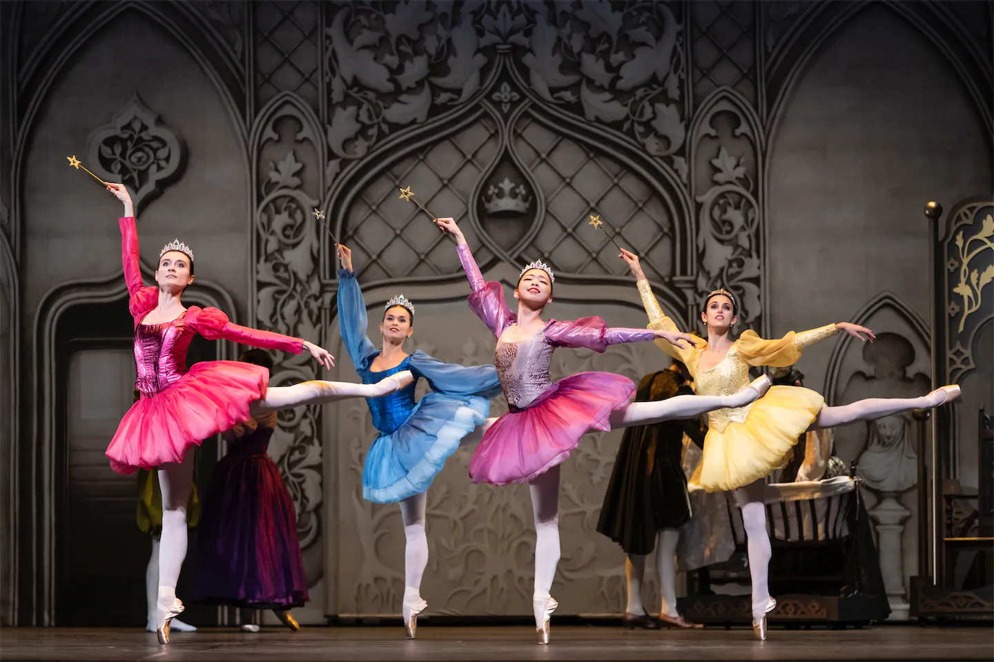 Quatre danseuses de ballet en tutus colorés roses, bleu, violet et jaune, portant des couronnes et tenant des baguettes étoilées, exécutant un grand écart sur scène devant un décor orné.