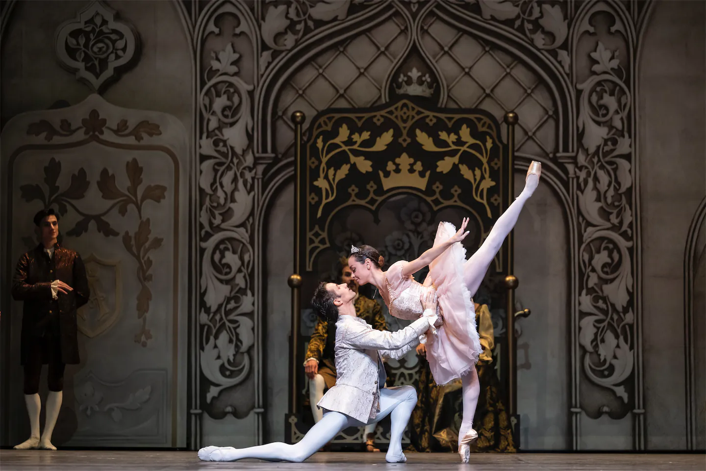 Deux danseurs de ballet effectuent une pose élégante devant un trône orné, l'homme en blanc agenouillé tenant la danseuse en tutu rose levant une jambe en pointe.