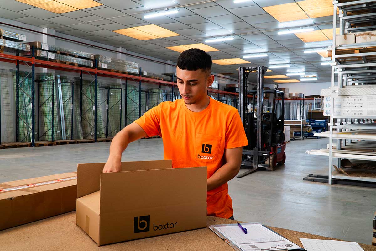 Empleado de almacén con camiseta naranja y logo 'boxtar' empacando una caja de cartón en una nave industrial.