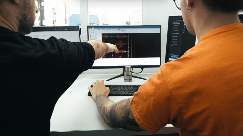 Dos hombres observando y trabajando en un diseño 3D en una computadora, uno señala la pantalla mientras el otro usa el teclado.