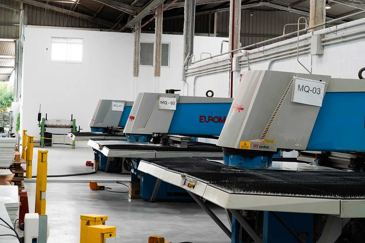 Tres máquinas industriales azules y grises etiquetadas como MQ-01, MQ-02 y MQ-03 en una fábrica moderna con paredes blancas.