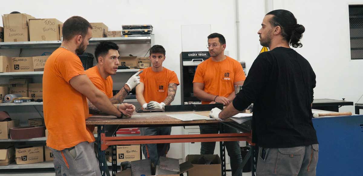 Cinco hombres reunidos en un taller industrial, cuatro con camiseta naranja y uno con camiseta negra, discutiendo alrededor de una mesa de trabajo.