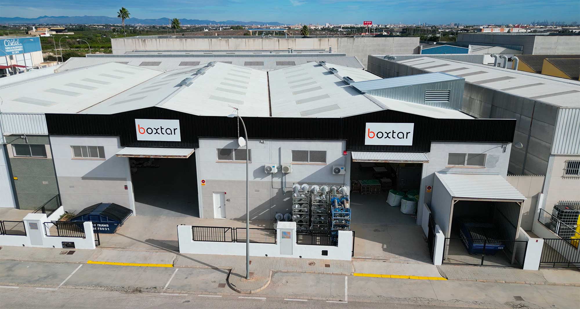 Nave industrial con dos entradas grandes y carteles con el nombre 'boxtar', ubicada en un polígono industrial bajo cielo azul.