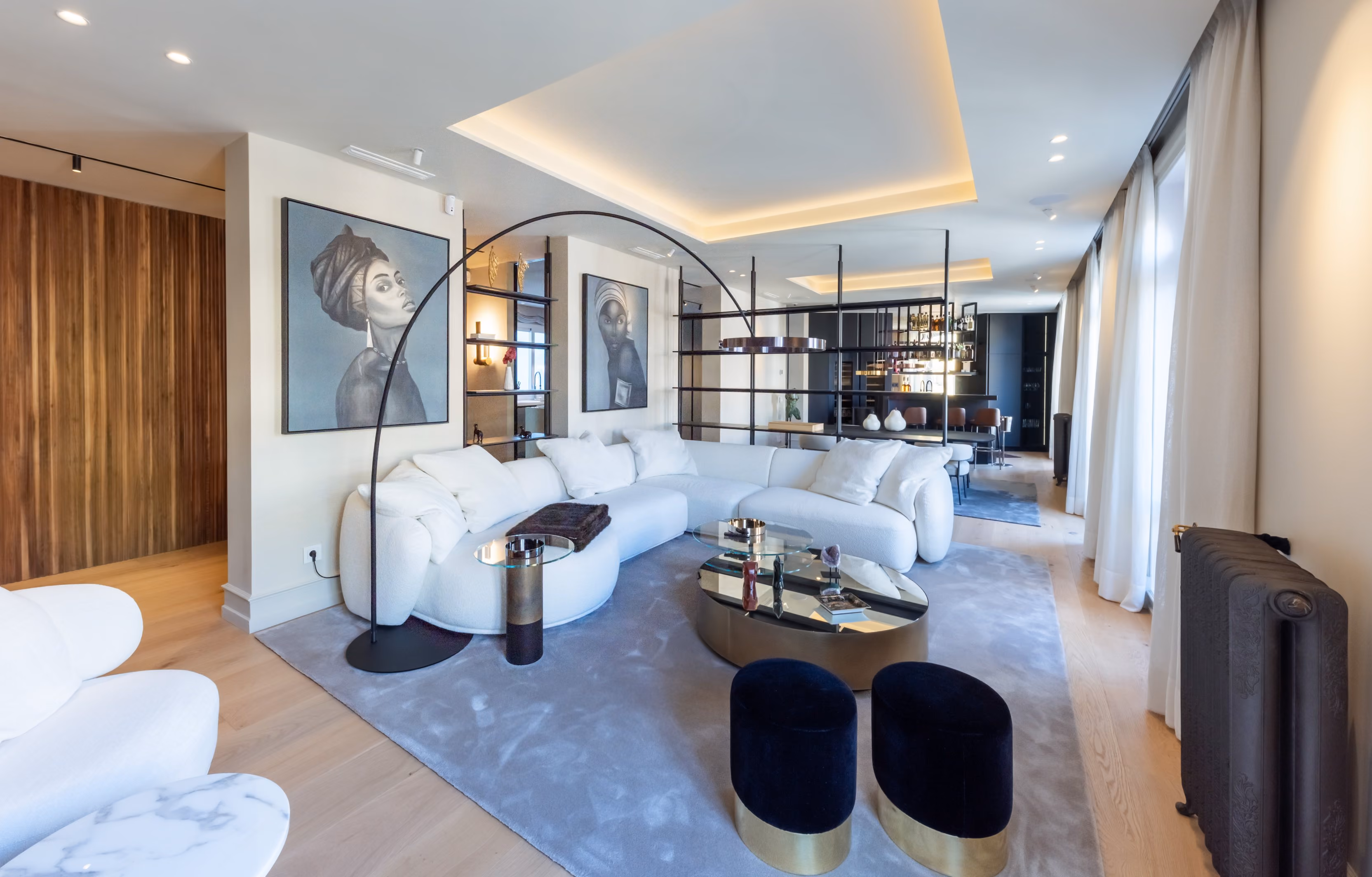 Salón contemporáneo y elegante con sofá modular blanco en forma de L, alfombra gris clara y mesas de centro doradas con superficies de espejo. Dos retratos artísticos en tonos monocromos decoran la pared, mientras una estantería abierta separa la zona de estar del comedor y la barra al fondo. La iluminación indirecta en el techo y la luz natural de los ventanales aportan calidez al espacio.