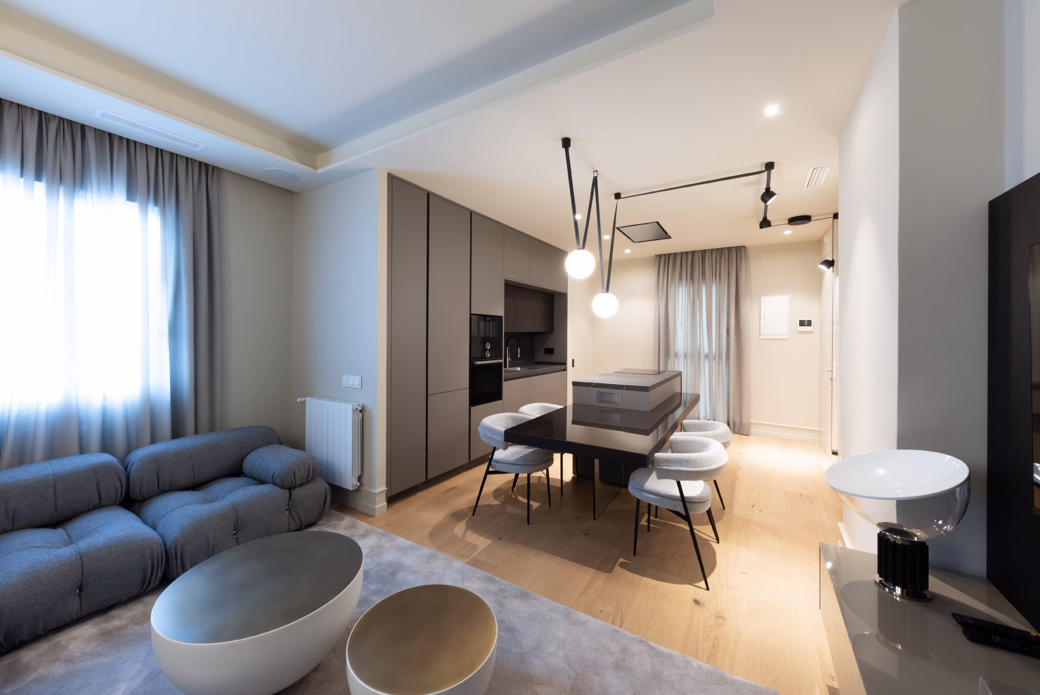 Apartamento moderno y acogedor con diseño de concepto abierto. A la izquierda, un sofá modular gris oscuro junto a una ventana con cortinas translúcidas. En el centro, una mesa de comedor rectangular con sillas tapizadas blancas y lámparas colgantes esféricas. Al fondo, una cocina de líneas minimalistas con muebles empotrados en tonos neutros y detalles en negro. El suelo de madera clara y la iluminación cálida completan la atmósfera elegante del espacio.