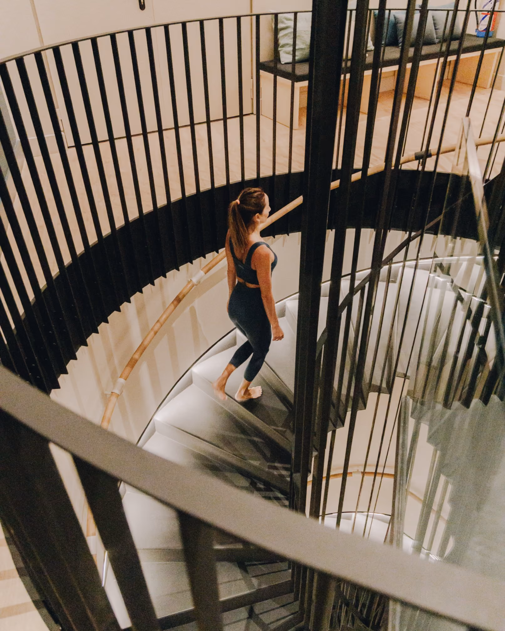 Mujer descalza subiendo una escalera de caracol moderna con peldaños grises y barandilla metálica negra. La escalera está iluminada sutilmente y se encuentra en un espacio interior de diseño contemporáneo, con banco de madera y cojines visibles en la planta superior.
