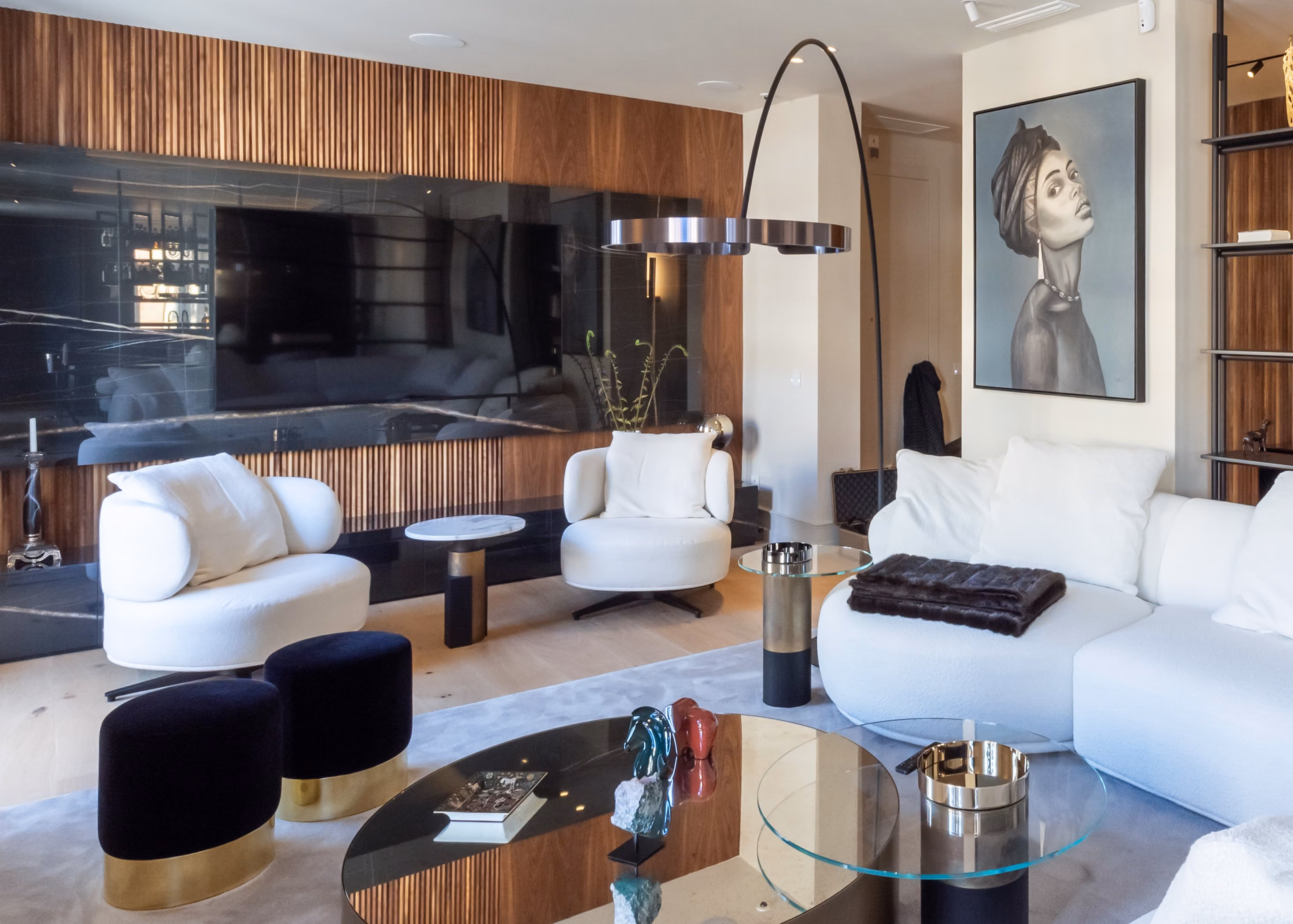 Salón contemporáneo y elegante con sofá modular blanco en forma de L, alfombra gris clara y mesas de centro doradas con superficies de espejo. Dos retratos artísticos en tonos monocromos decoran la pared, mientras una estantería abierta separa la zona de estar del comedor y la barra al fondo. La iluminación indirecta en el techo y la luz natural de los ventanales aportan calidez al espacio.