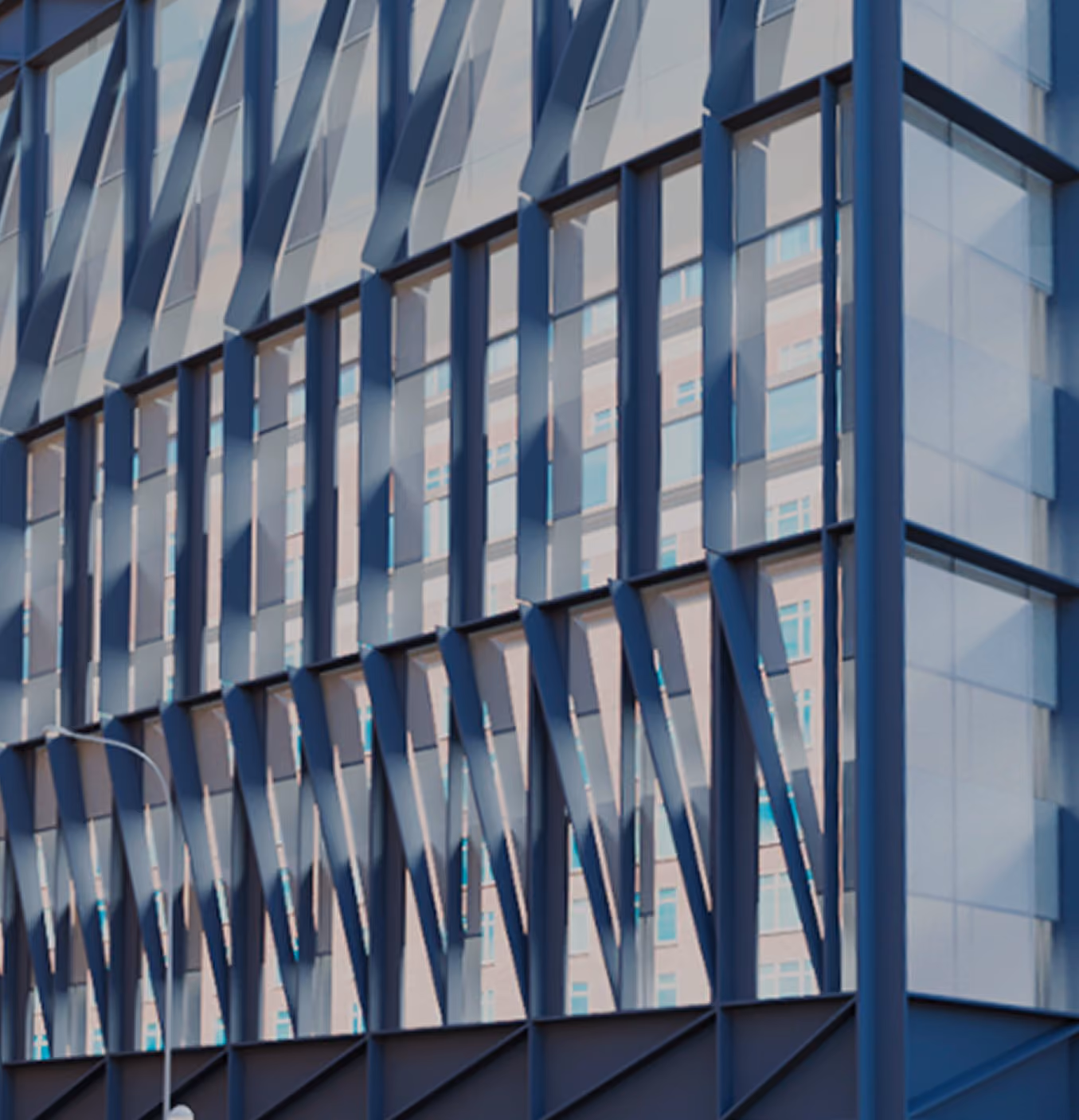 Edificio institucional de fachada acristalada con estructura metálica diagonal azul oscuro, ubicado en una esquina urbana. En la parte superior derecha de la fachada se observa un logotipo rojo con la letra 'R'. El entorno incluye edificios residenciales adyacentes, árboles frondosos y una calle despejada bajo cielo azul.