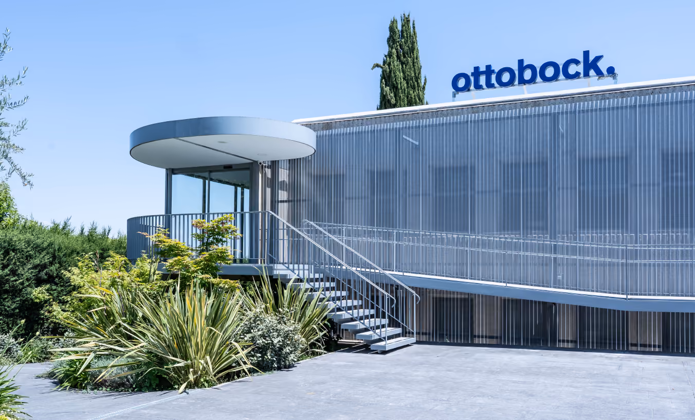 Fachada exterior del edificio de Ottobock, con revestimiento translúcido de policarbonato ondulado y acceso elevado mediante escaleras y rampa metálica. A la izquierda, una estructura cilíndrica con cubierta circular protege la entrada acristalada. El cielo despejado y los cipreses de fondo enmarcan el edificio con un entorno natural.