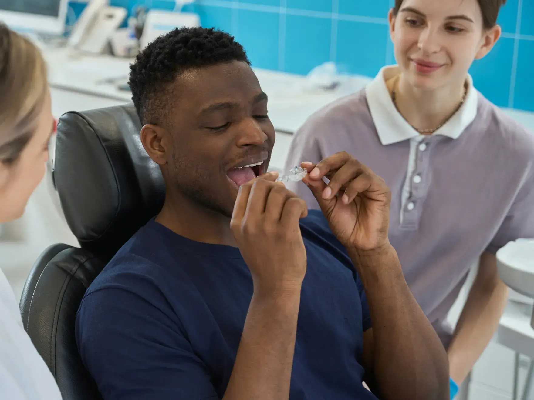 A man in a dental chair is trying on a clear aligner while two people observe.