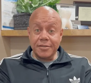 Man in a tracksuit jacket is speaking directly to the camera indoors.