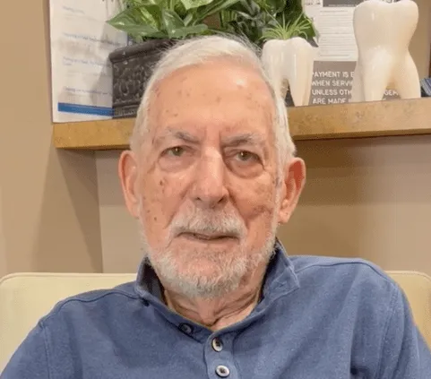 An older man with a white beard wearing a blue shirt sits indoors, looking at the camera.