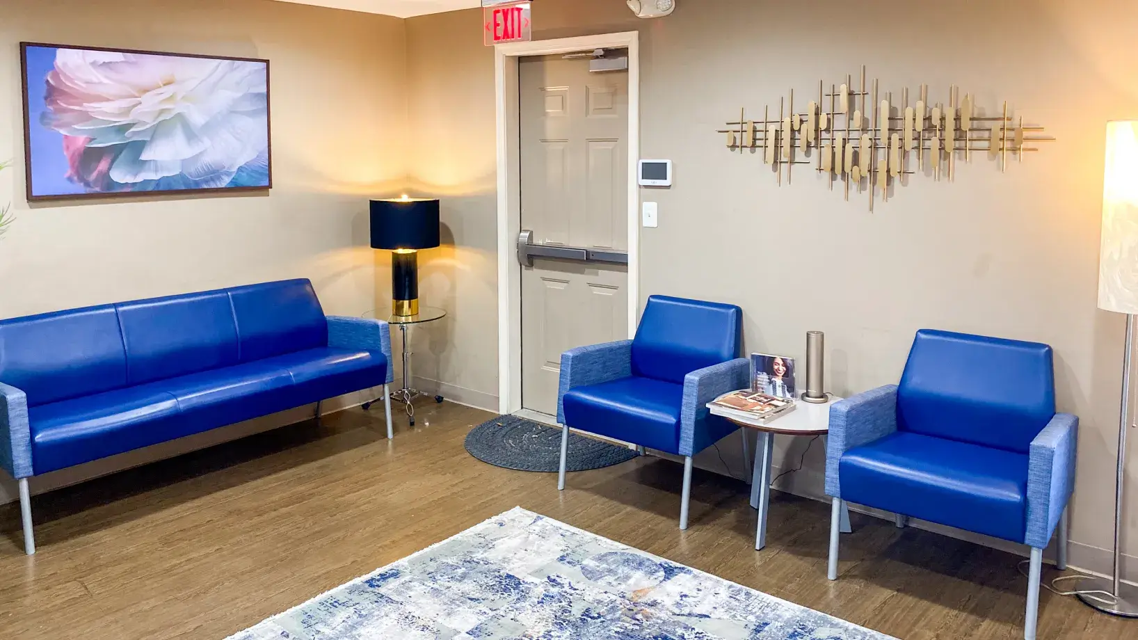 A waiting room with blue seating, a lamp, wall art, and magazines on a small table.