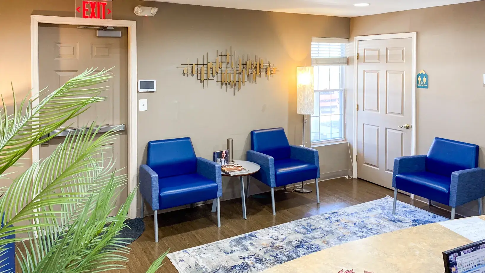 Small waiting room with three blue chairs, a table, and an exit sign above the door.