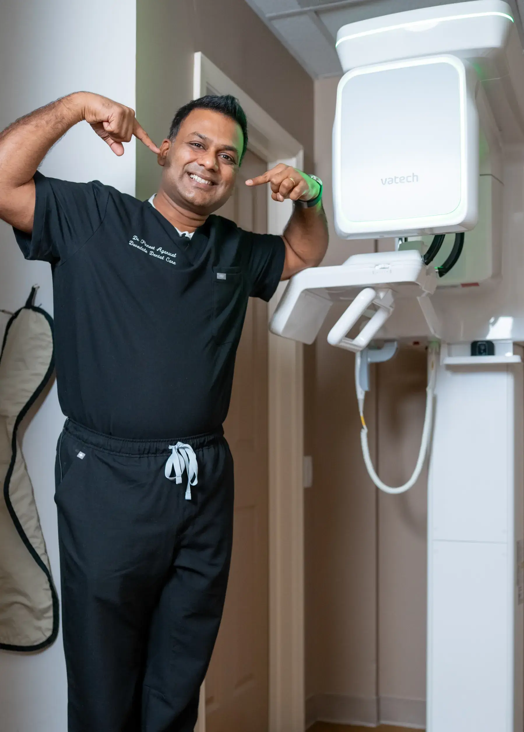 A dentist talks to a seated patient while an assistant works nearby in a dental office.