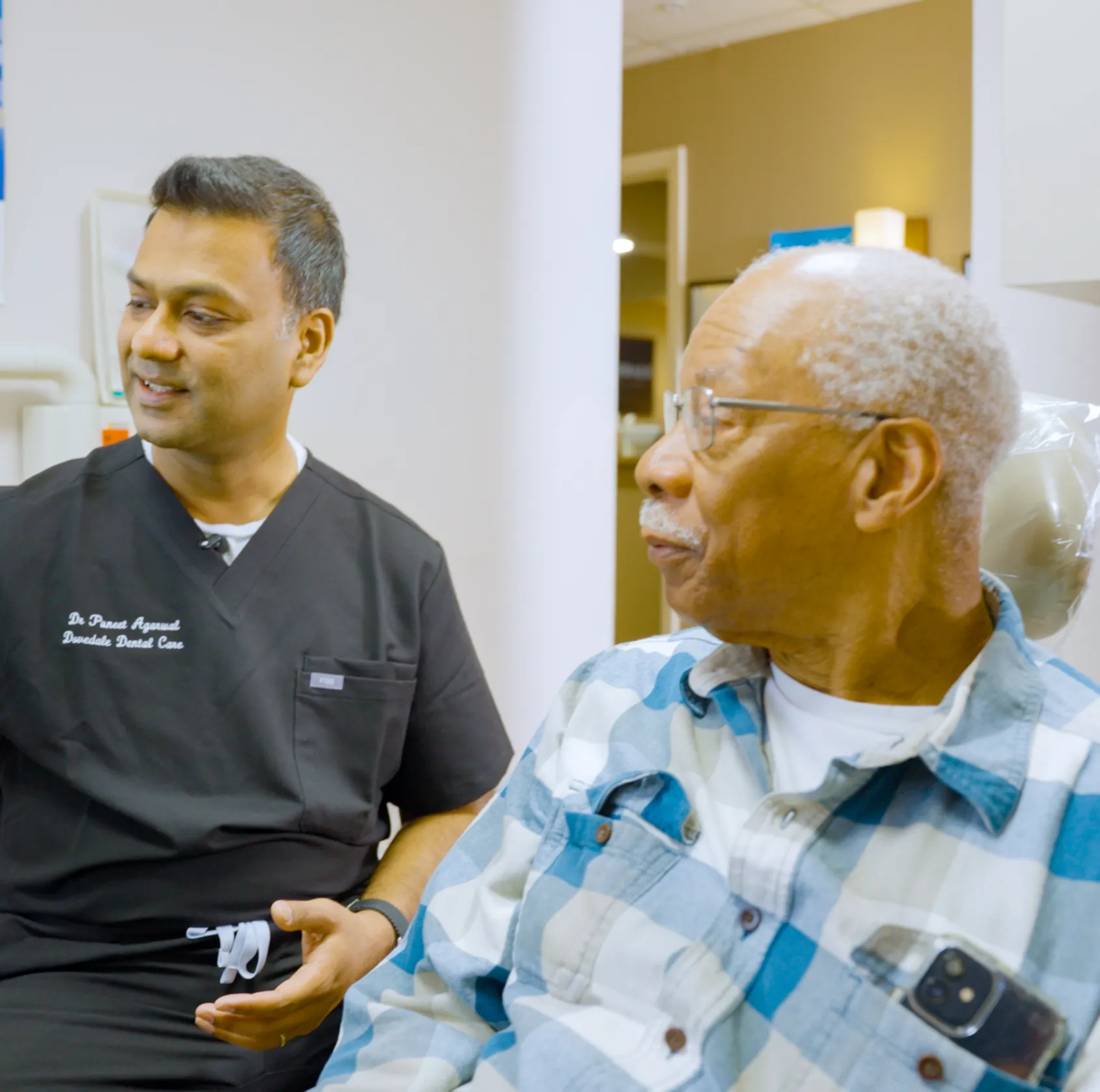 A dentist in black scrubs talks to an older man in a plaid shirt seated in a dental chair.