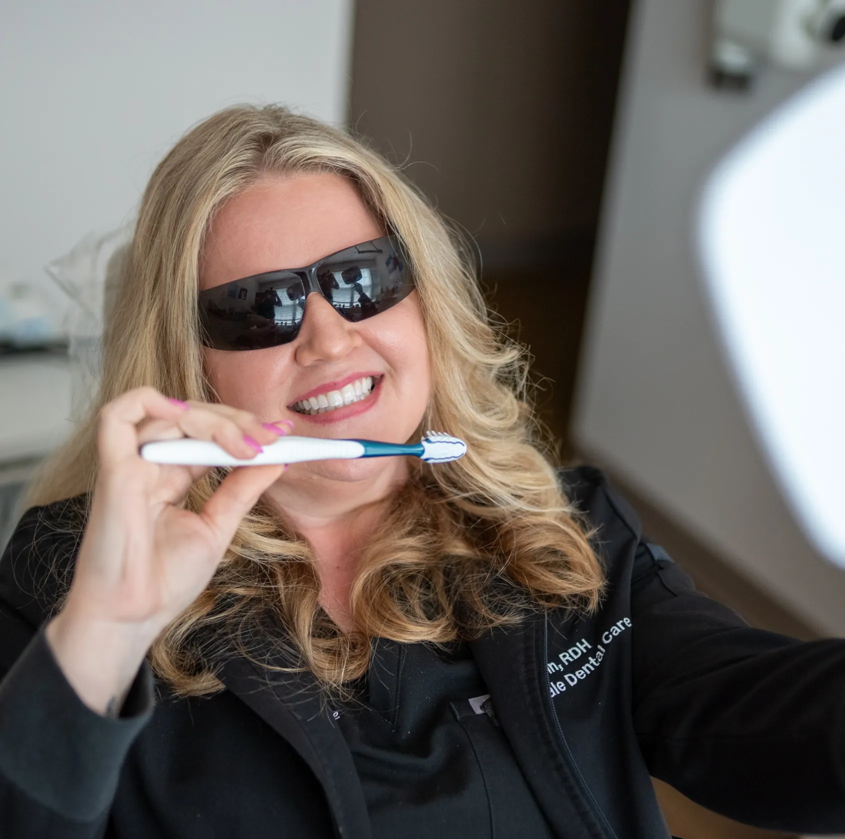 A woman wearing sunglasses holds a toothbrush while smiling and looking at a mirror.
