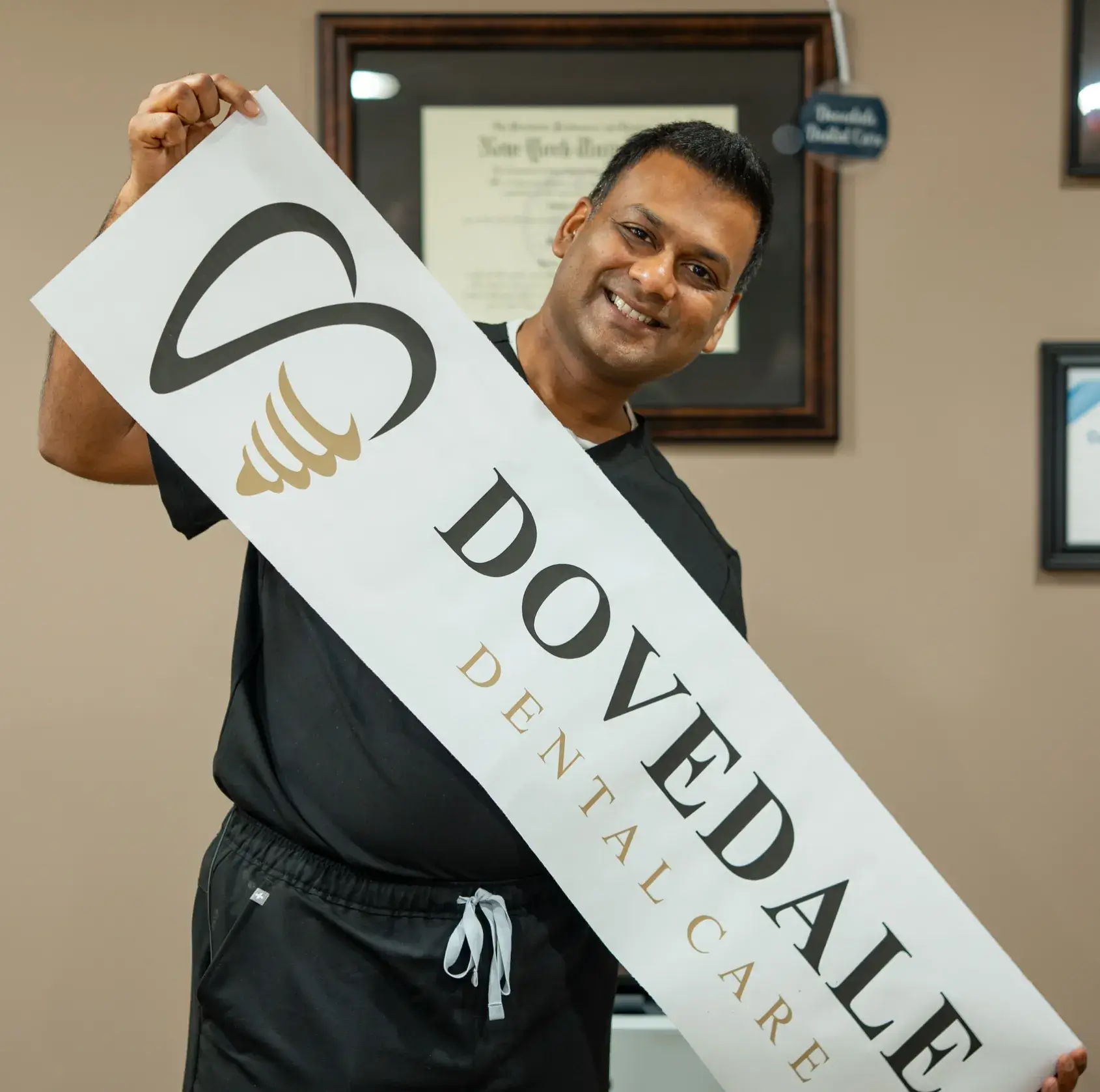 A smiling man in a black dental uniform points to his head with both hands.