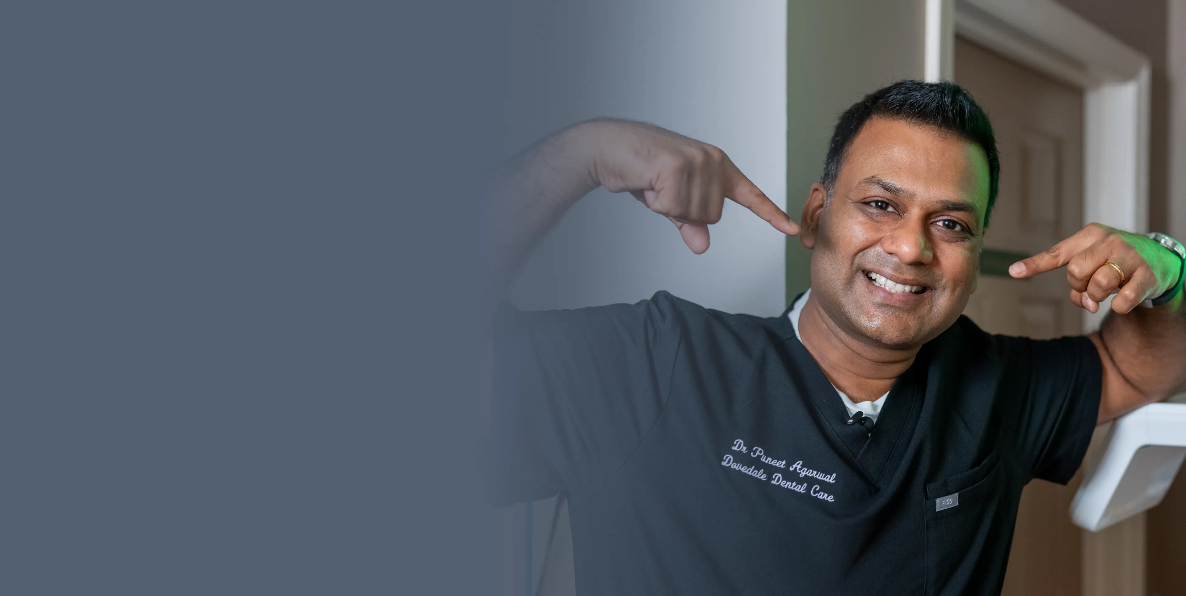 A smiling person in a dental uniform poses, pointing to their teeth.