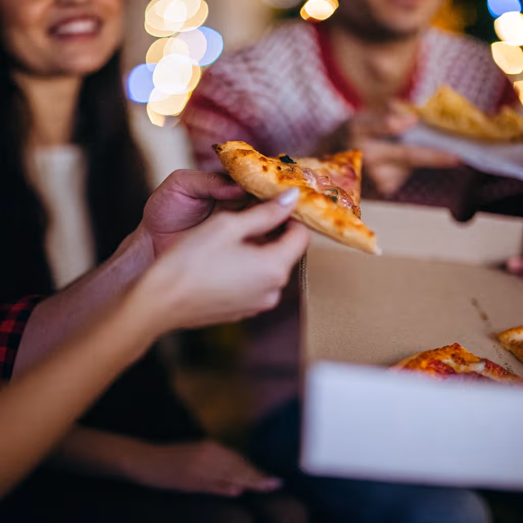People enjoying pizza slices from an open takeaway pizza box with blurred festive lights in the background.