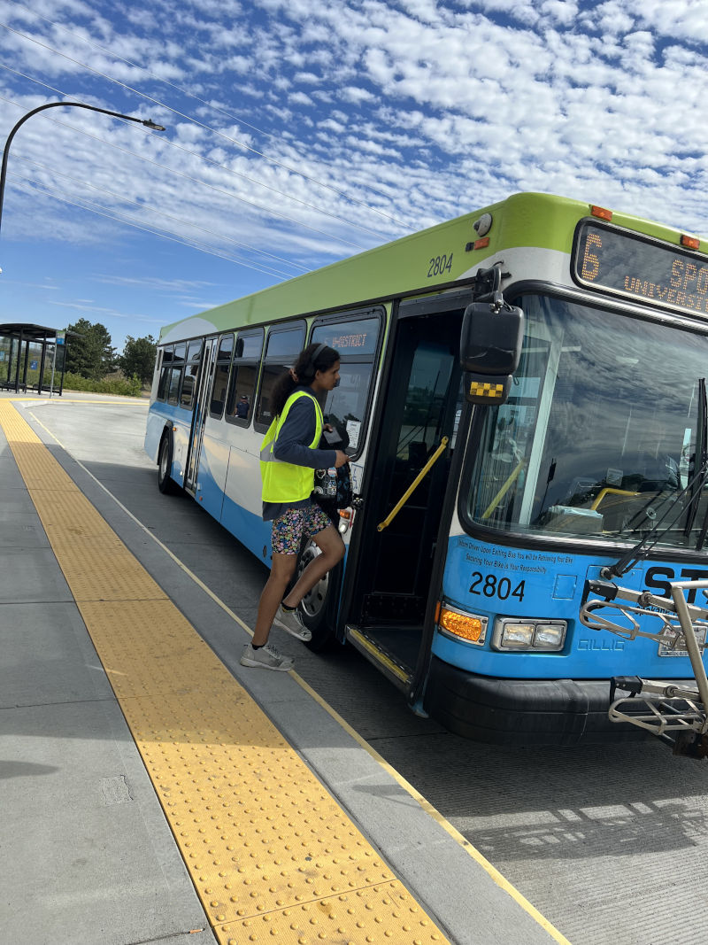 A person wearing a yellow safety vest boards a city bus at a transit station on a sunny day with scattered clouds. The bus displays the number 6 and has blue and green colors.