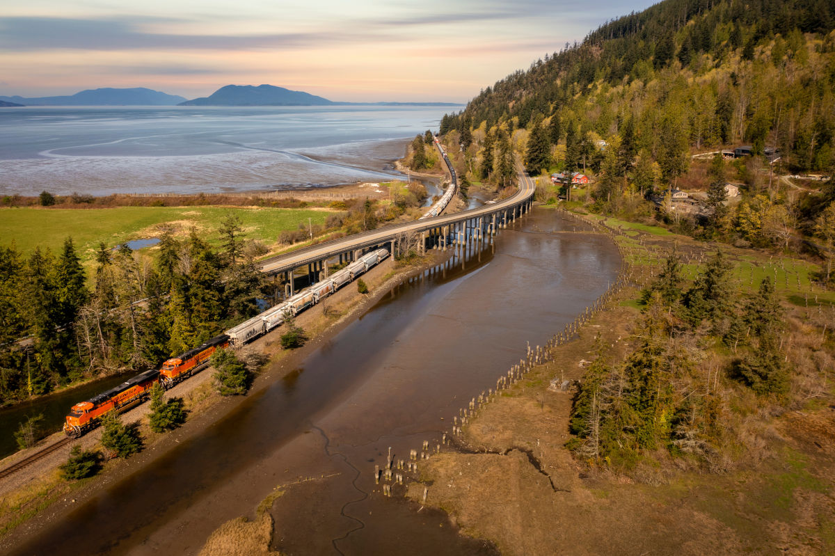 Aerial view of a freight train passing through a lush landscape with a highway overpass, coastal wetlands, tall trees, and distant mountains under a colorful sky near sunset