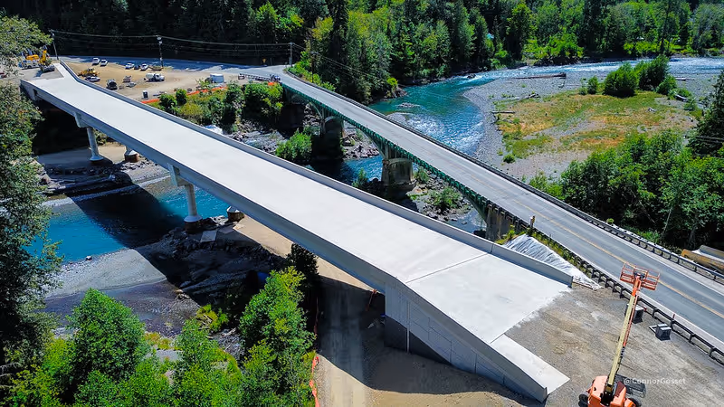 Aerial view of a newly constructed concrete bridge spanning a river alongside an older bridge. The scene is surrounded by lush green trees, with construction vehicles and equipment visible near the bridge approach.