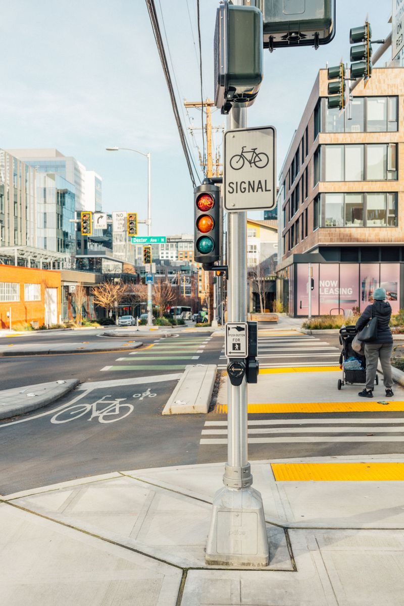 A city intersection in Downtown Seattle with a bike signal, a bike lane, and a crosswalk. A person pushes a stroller across the street, and nearby buildings are visible under a clear sky.