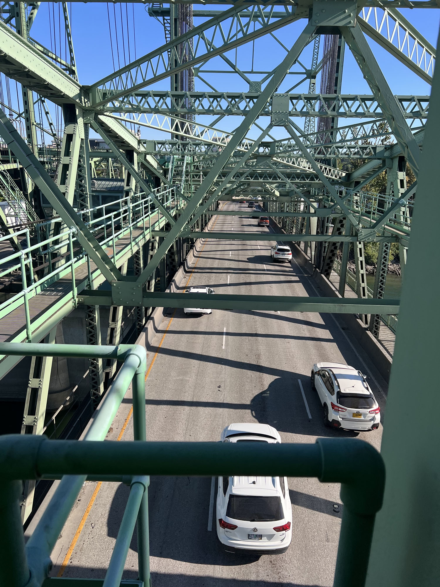 A shot looking down on a bridge through the green metal support beams on a clear day. Multiple cars drive across the bridge.